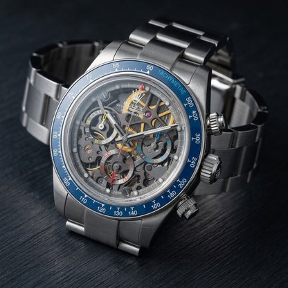 Steel skeleton chronograph at a three-quarter angle on dark slate, the fluted crown and pushers beside the blue bezel