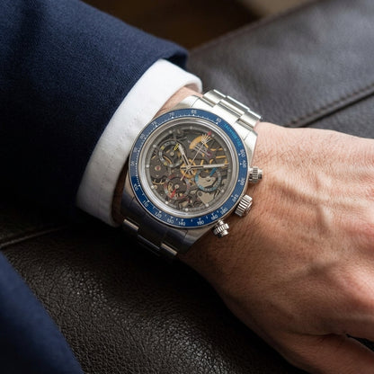 Steel skeleton chronograph with a blue bezel on its steel bracelet, worn on the wrist below a dark suit cuff