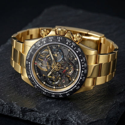Yellow gold skeleton chronograph at a three-quarter angle on dark slate, the gold pushers beside the black tachymetre bezel