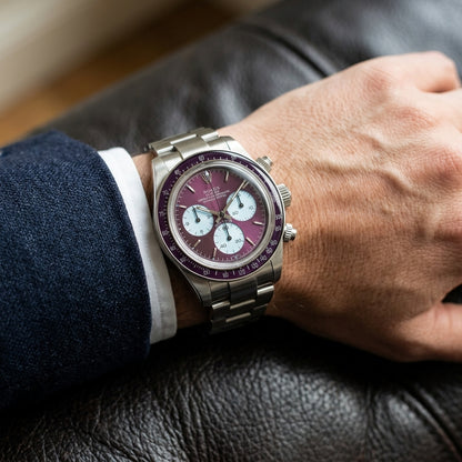 Steel chronograph with a plum dial on its steel bracelet, worn on the wrist below a dark suit cuff