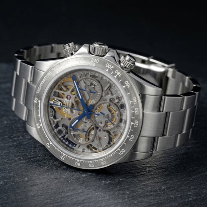 Steel skeleton chronograph at a three-quarter angle on dark slate, the clear tachymetre bezel beside the fluted crown and pushers
