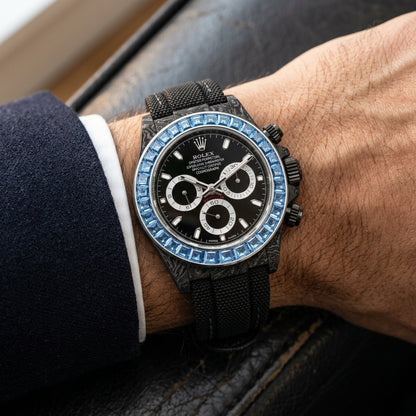 Black forged-carbon chronograph with a sky-blue stone bezel on its black woven strap, worn on the wrist below a dark suit cuff