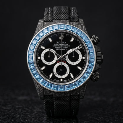 Black forged-carbon chronograph with a sky-blue stone bezel on its black woven strap, worn on the wrist below a dark suit cuff