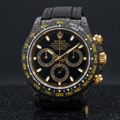Black forged carbon chronograph with a black dial printed in gold and a yellow tachymeter scale, upright on polished black granite