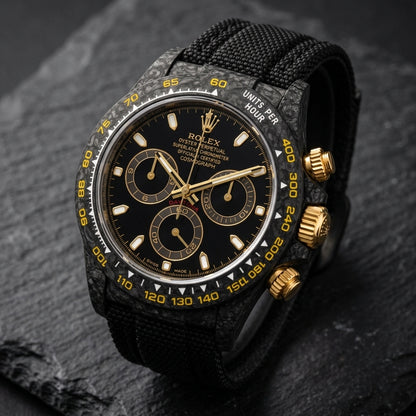 Black forged carbon chronograph at a three-quarter angle on dark slate, its gold crown and pushers against the marbled carbon flank