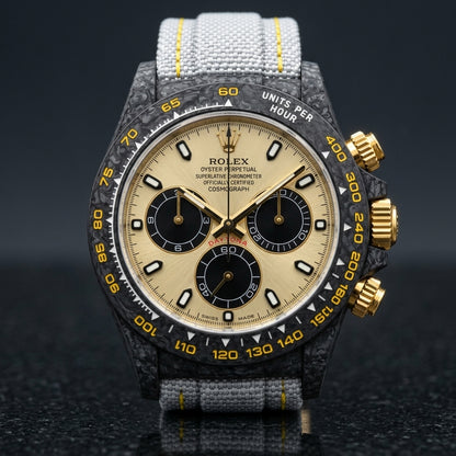 Black carbon chronograph with a champagne dial on its light-grey woven strap with yellow stitching, worn on the wrist below a suit cuff
