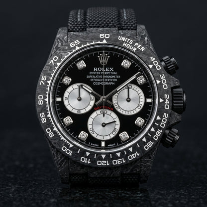 Black forged carbon chronograph with a black dial, silver counters and clear stone hour markers, upright on polished black granite