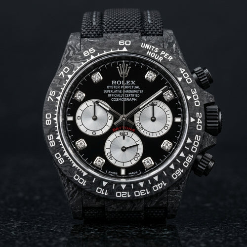 Black forged carbon chronograph with a black dial, silver counters and clear stone hour markers, upright on polished black granite