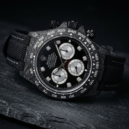 Black forged carbon chronograph at a three-quarter angle on dark slate, its black crown and pushers against the marbled carbon flank