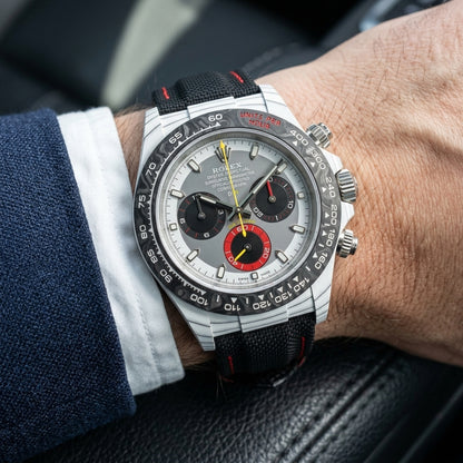 White carbon chronograph on its black canvas strap with red stitching, worn on the wrist below a navy suit cuff