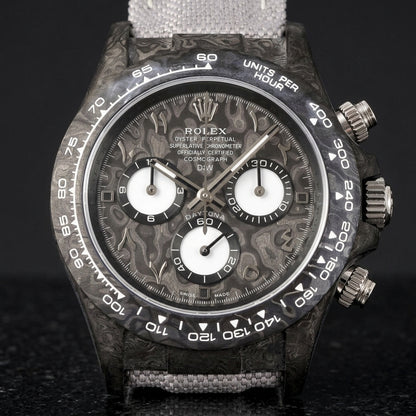 Black forged-carbon chronograph with a grey carbon dial on its grey canvas strap, worn on the wrist below a dark suit cuff