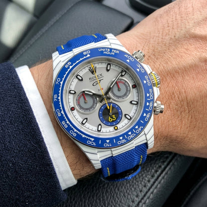 White forged-carbon chronograph with a blue bezel on its blue canvas strap, worn on the wrist below a dark suit cuff