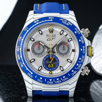 White forged-carbon chronograph with a blue bezel on its blue canvas strap, worn on the wrist below a dark suit cuff
