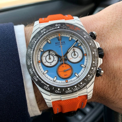 White carbon chronograph with a sky-blue dial on its orange canvas strap, worn on the wrist below a dark suit cuff
