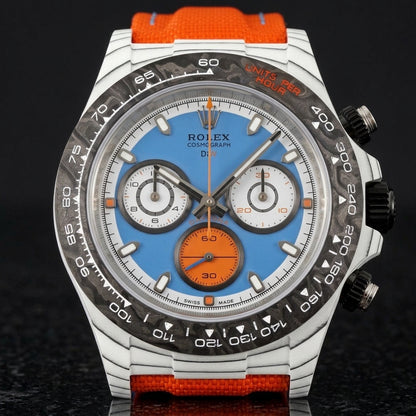White carbon chronograph with a sky-blue dial on its orange canvas strap, worn on the wrist below a dark suit cuff