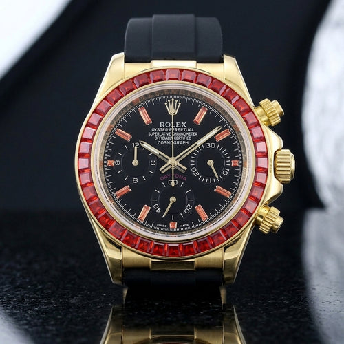 Gold-tone chronograph with a red baguette-stone bezel and a black dial on a black rubber strap, upright on polished black granite