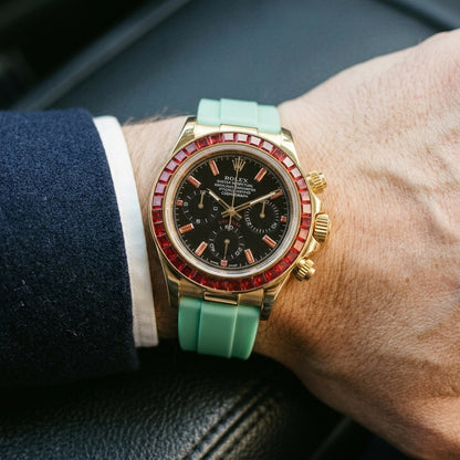 Gold chronograph with a red stone bezel on its mint-green rubber strap, worn on the wrist below a dark suit cuff