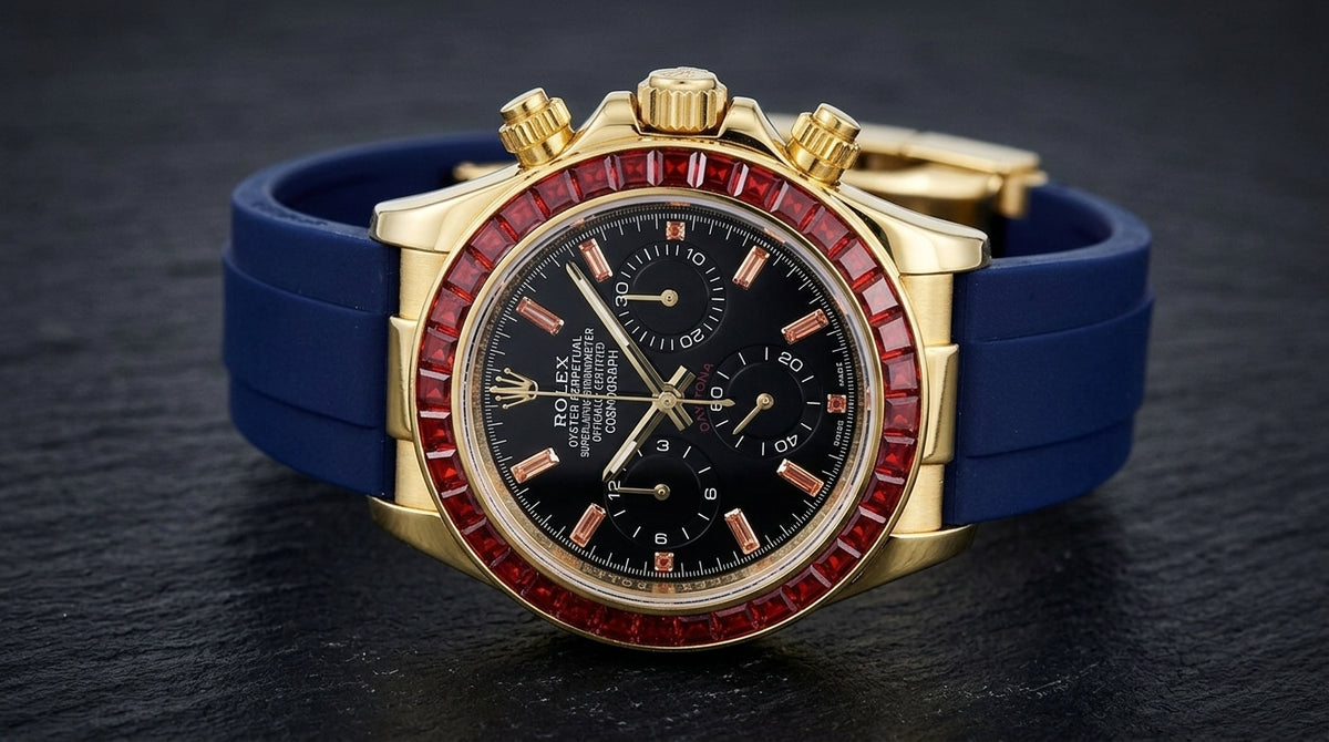 Gold chronograph at a three-quarter angle on dark slate, the fluted crown and pushers beside the ring of red baguette-cut stones