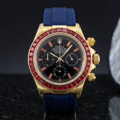 Gold chronograph with a red stone bezel on its navy blue rubber strap, worn on the wrist below a dark suit cuff