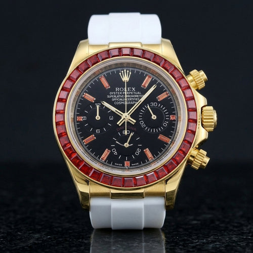 Gold-tone chronograph with a red baguette-stone bezel and a black dial on a white rubber strap, upright on polished black granite