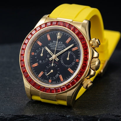 Gold-tone chronograph at a three-quarter angle on dark slate, the fluted crown and pushers beside the red stone bezel and the yellow rubber strap