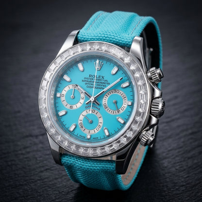 Steel chronograph at a three-quarter angle on dark slate, the clear bezel stones beside the fluted steel crown and pushers and the turquoise canvas strap curving away