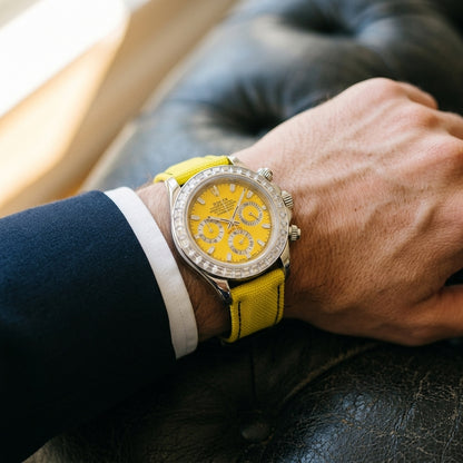 Steel chronograph with a yellow dial on its lemon-yellow canvas strap, worn on the wrist below a dark suit cuff