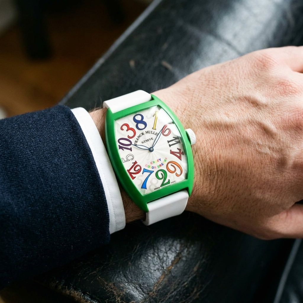Green curved tonneau watch with rainbow numerals on its white rubber strap, worn on the wrist below a dark suit cuff