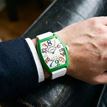 Green curved tonneau watch with rainbow numerals on its white rubber strap, worn on the wrist below a dark suit cuff