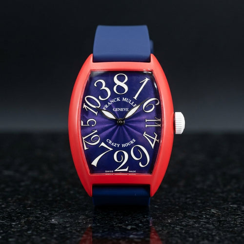 Red curved tonneau watch with a blue dial and white numerals set out of order, upright on polished black granite