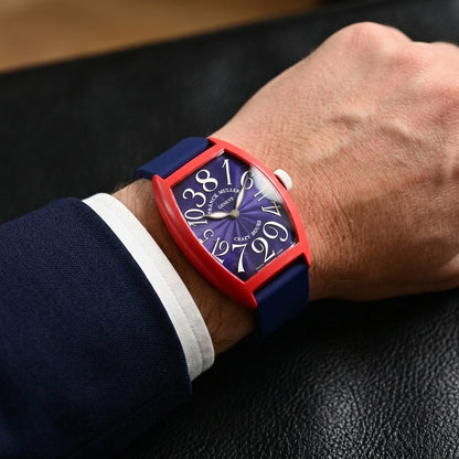 Red curved tonneau watch on its navy rubber strap, worn on the wrist below a dark suit cuff