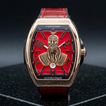 Rose gold curved tonneau watch with a red dial on its red strap, worn on the wrist below a dark suit cuff