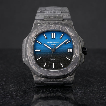 Forged carbon sports watch with a blue-to-black gradient dial and white baton markers, upright on polished black granite