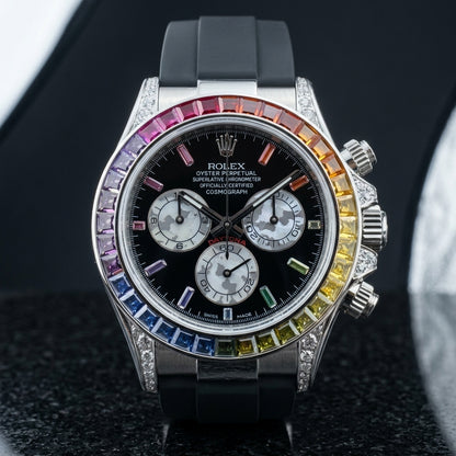 Steel chronograph with a rainbow stone bezel on a black rubber strap, on the wrist below a dark suit cuff