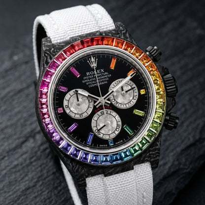 Black forged carbon chronograph at a three-quarter angle on dark slate, its black crown and pushers beside the rainbow bezel