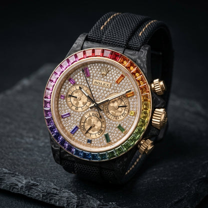 Black forged carbon chronograph at a three-quarter angle on dark slate, its gold crown and pushers beside the rainbow bezel