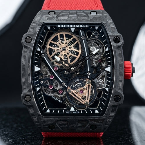Black carbon skeleton tonneau watch with a red crown and red woven strap, upright on polished black granite