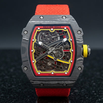 Carbon tonneau skeleton watch with a red flange and yellow track on a red elastic strap, upright on polished black granite