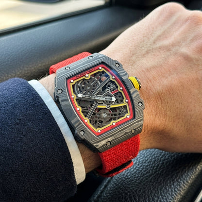 Carbon tonneau skeleton watch on its red elastic strap, worn on the wrist below a dark suit cuff