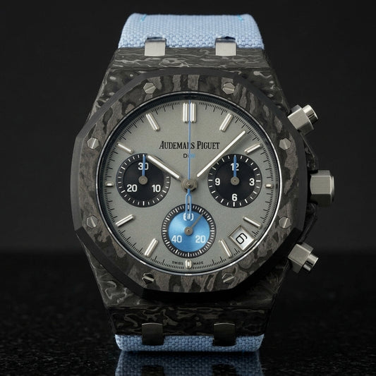 Black forged-carbon octagonal chronograph with a grey dial and a sky-blue counter, upright on polished black granite