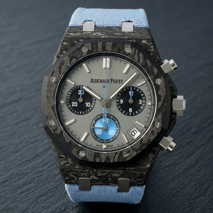 Black forged-carbon octagonal chronograph at a three-quarter angle on dark slate, whitish carbon veining on its flank beside the steel crown and pushers
