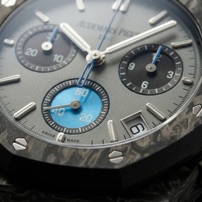 Macro of the grey dial with steel batons, two black counters and the sky-blue counter at six inside the octagonal carbon bezel