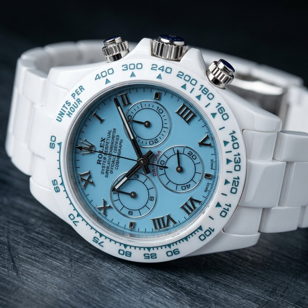 White ceramic chronograph at a three-quarter angle, the teal-blue tachymeter bezel beside the steel crown and pushers with blue cabochons