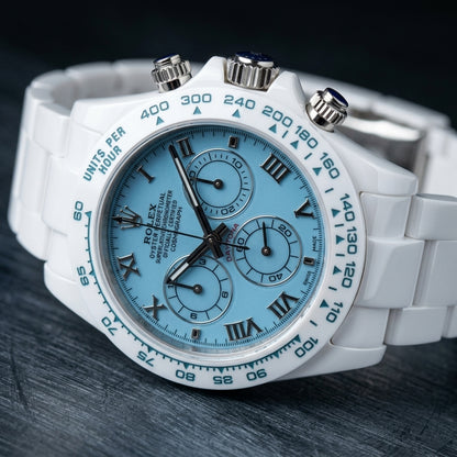 White ceramic chronograph at a three-quarter angle, the teal-blue tachymeter bezel beside the steel crown and pushers with blue cabochons