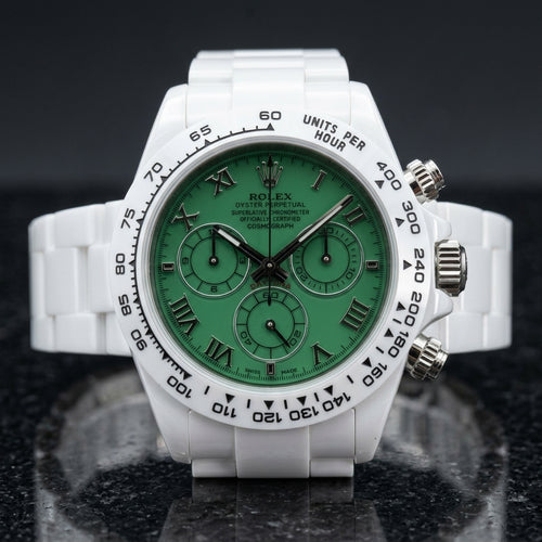 White ceramic chronograph with a green Roman numeral dial and a white tachymeter bezel, upright on polished black granite