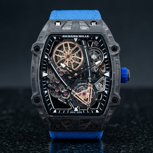 Black carbon tonneau watch with an open skeleton dial and a blue woven strap, upright on polished black granite