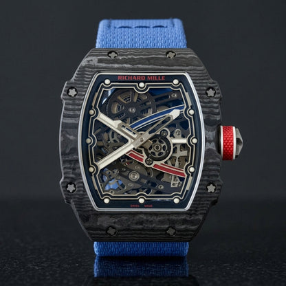 Ultra-thin black carbon tonneau watch with a blue and red skeleton dial and a blue textile strap, upright on polished black granite