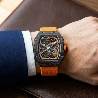 Black carbon tonneau watch with orange details on an orange textile strap, worn on the wrist below a navy suit cuff