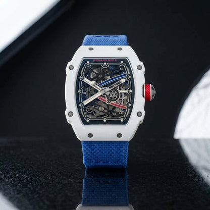 White tonneau watch with a red crown on a blue textile strap, worn on the wrist below a navy suit cuff
